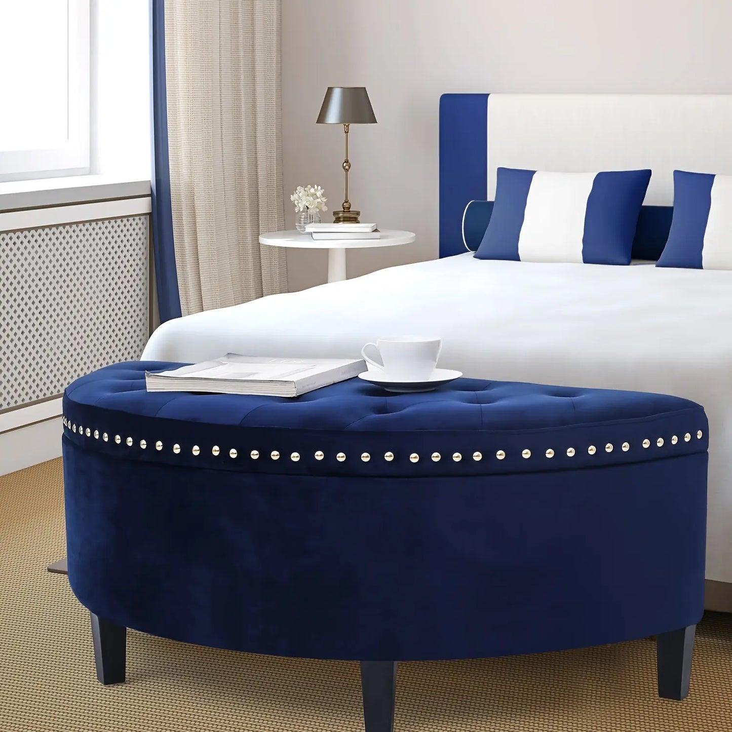 The Rhythm Velvet Storage Ottoman by Fantascticz, shown in navy blue as a sophisticated bench at the foot of a bed. The elegant button-tufted top and silver nailhead trim make it a luxurious addition to any bedroom.