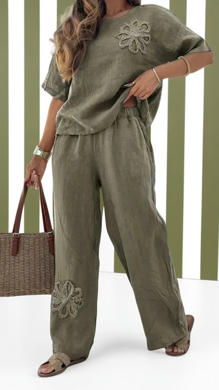 Woman wearing an olive green outfit with floral embroidery, holding a woven bag against a striped background.