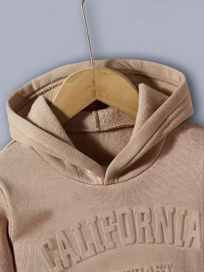 A close-up view of a tan children’s hoodie on a wooden hanger, showcasing the high-quality embossed ‘CALIFORNIA’ lettering across the chest and the soft fleece texture of the fabric.