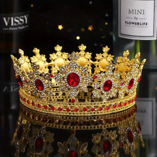 Styled photo of The Sovereign Crown, featuring a gold-tone baroque design with large ruby-red jewels and crystal accents. The crown rests on a black reflective surface, creating a stunning mirror image.