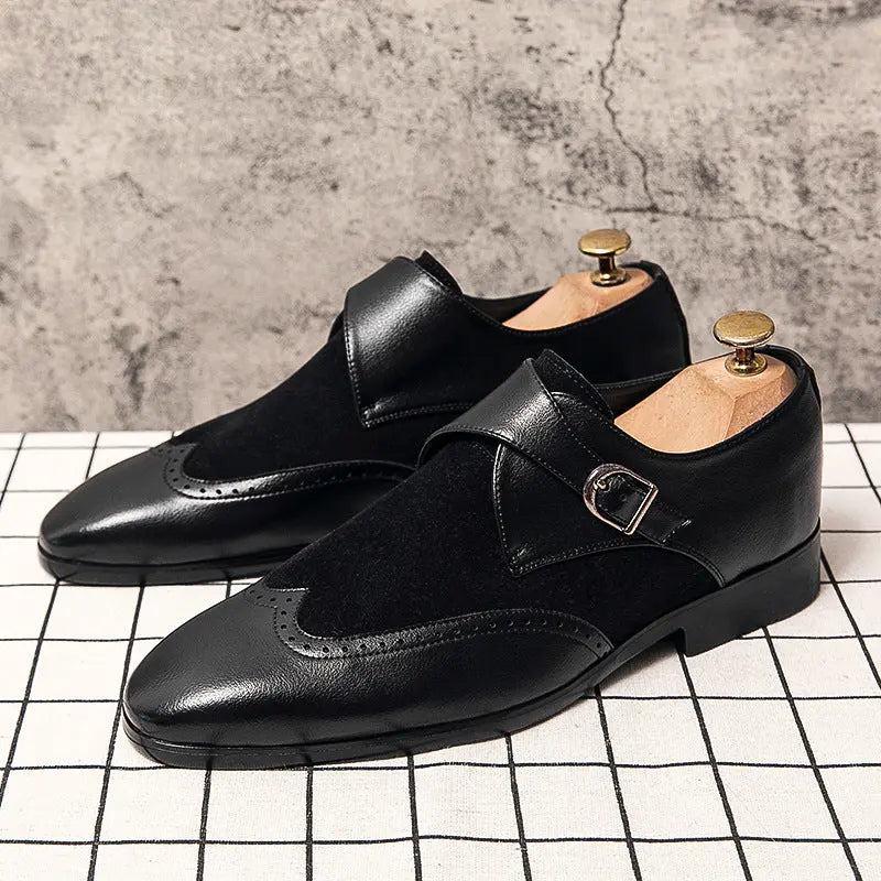 Product view of The Sterling Monk Strap shoes in all-black. The shoes feature a mix of smooth leather and suede, brogue detailing, and are displayed on a white grid-tile surface against a concrete wall.